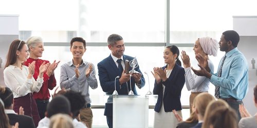 businessman-holding-award-on-podium-with-colleague-D5CQW3R-min.jpg